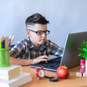 A young boy wearing glasses focused on using a laptop for online learning, surrounded by school supplies.